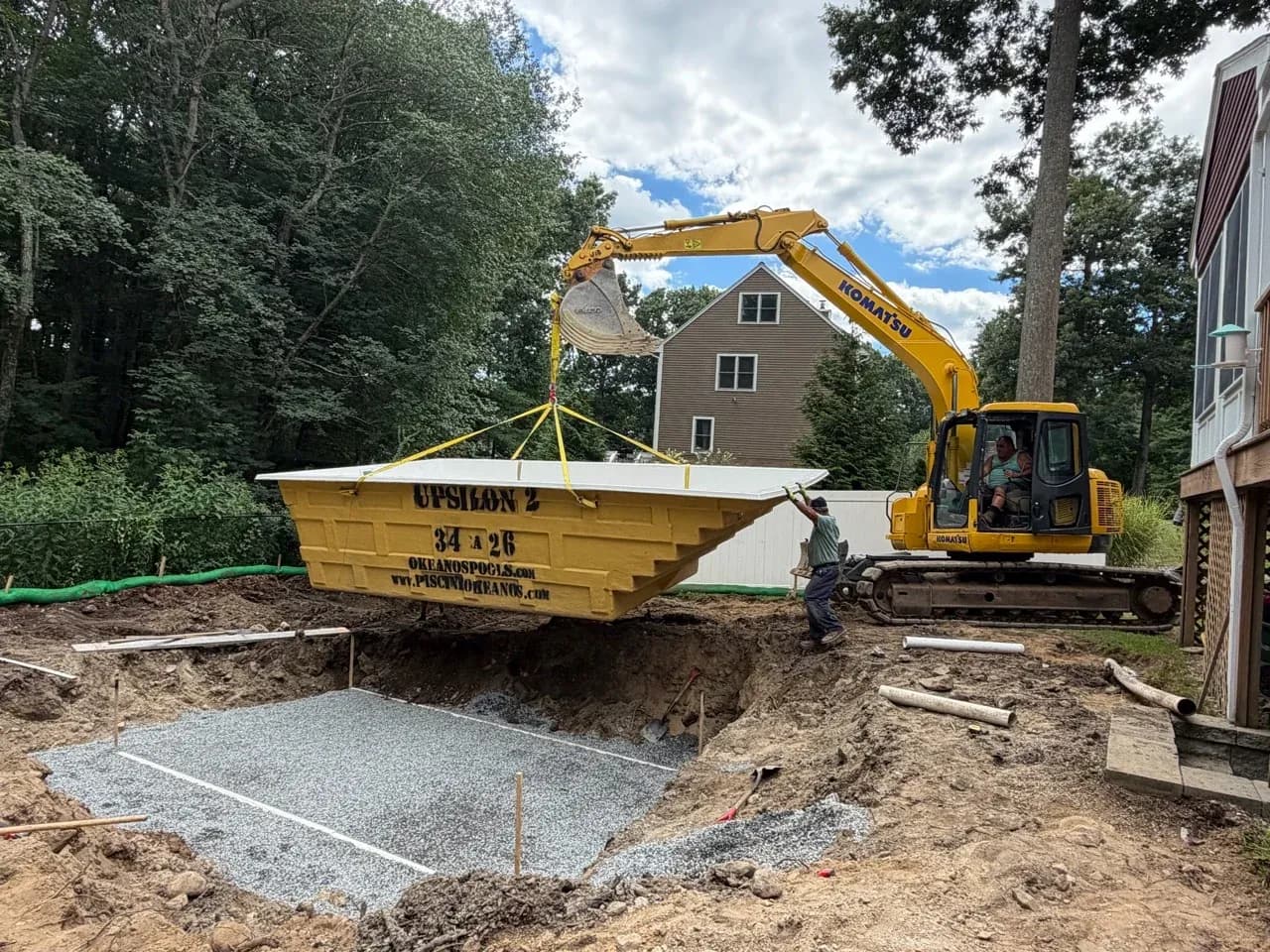 Crew guiding an Okeanos fiberglass pool shell over a gravel-lined excavation.