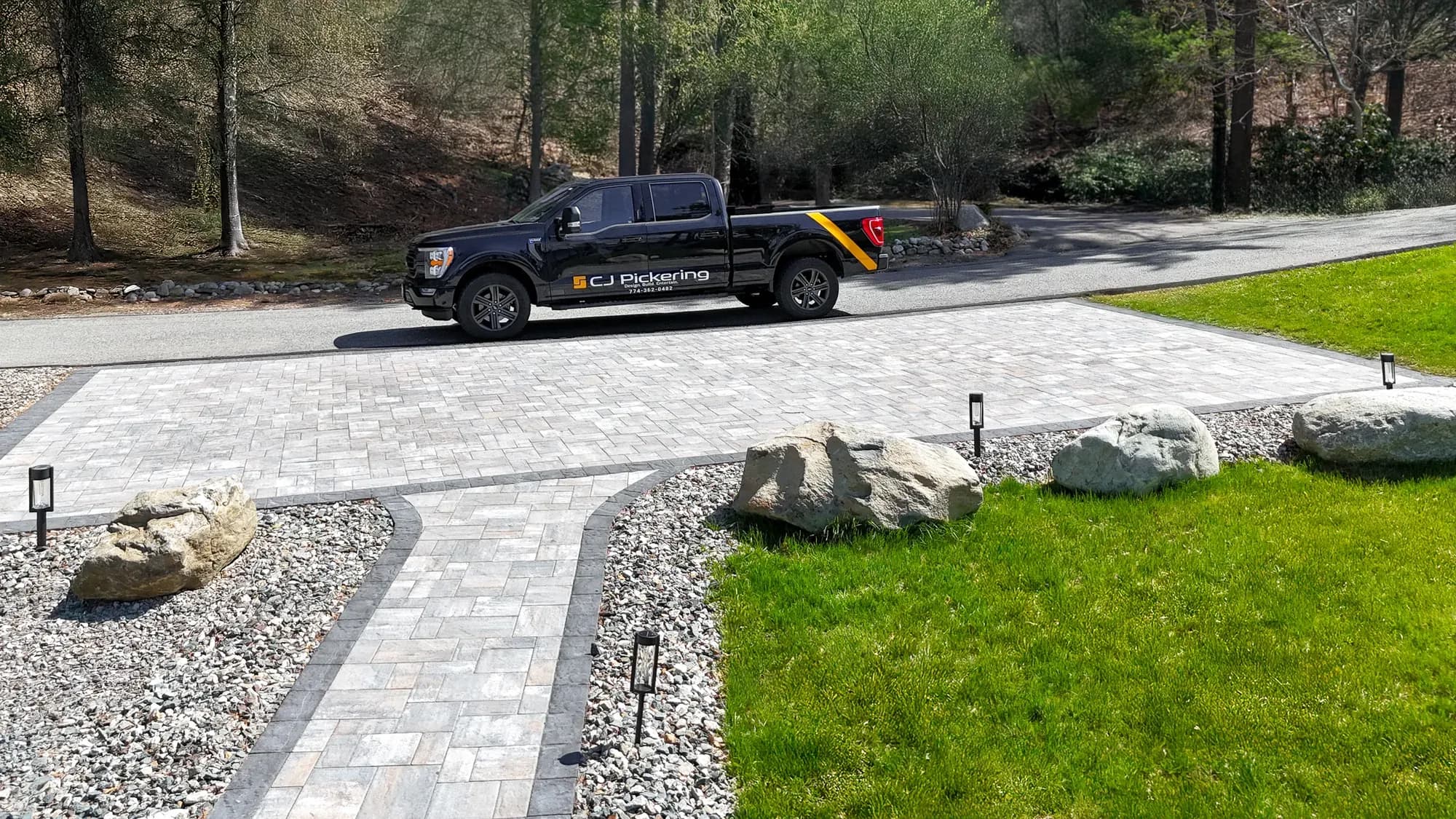Curved paver entry walkway, path lights, gravel beds, and boulders beside the upper driveway.