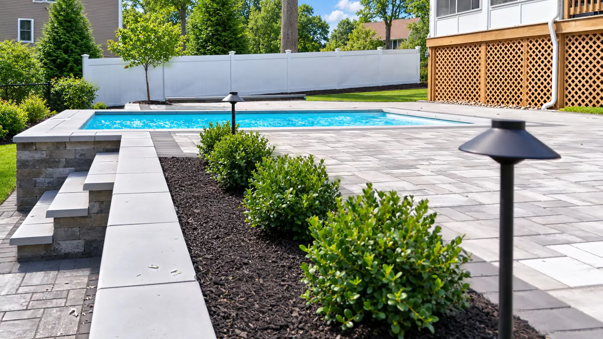 Capped retaining wall, raised planting bed, and path lights beside the pool patio in Mansfield, MA.