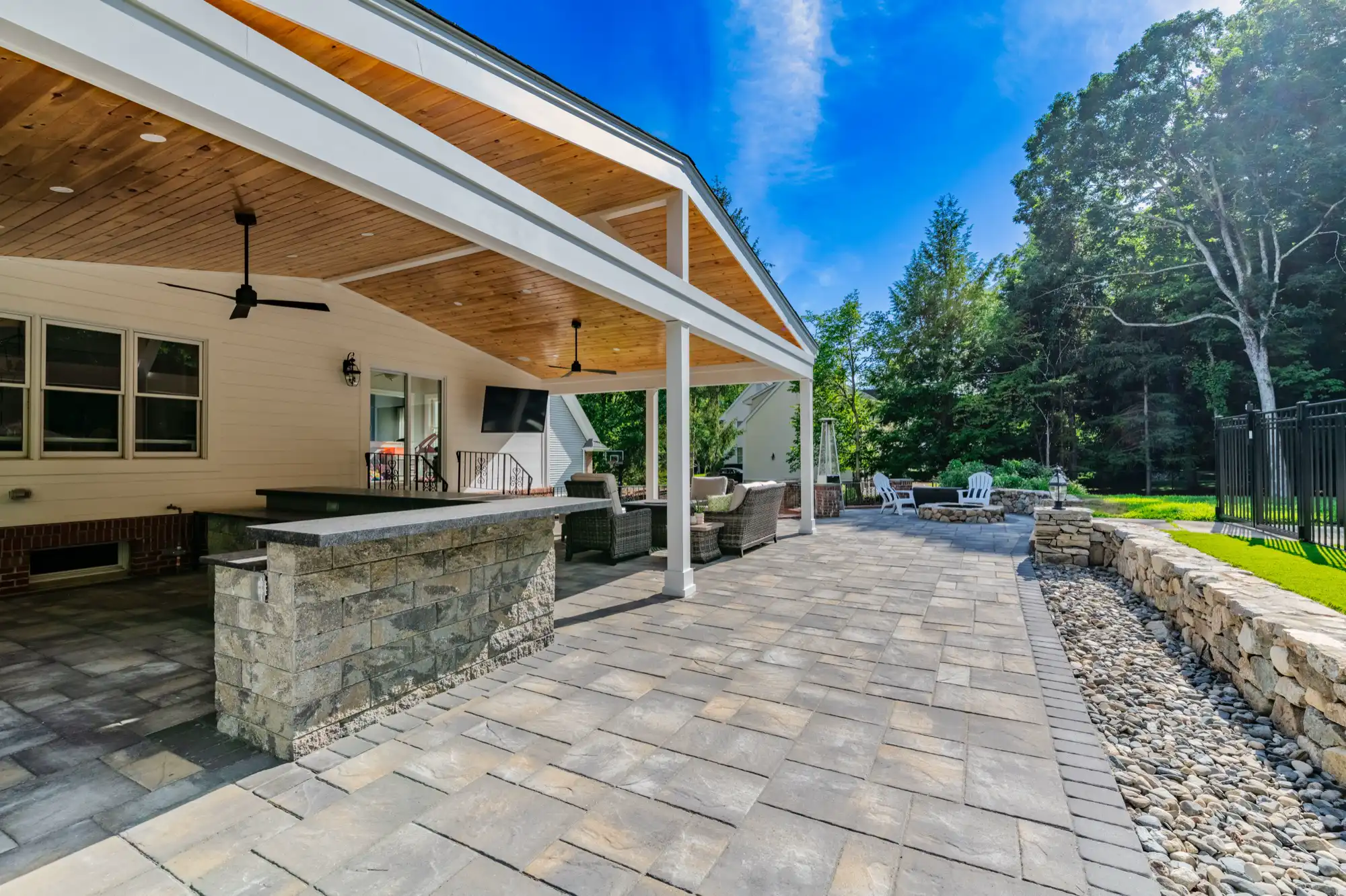Outdoor kitchen and broad paver terrace beneath and beside the wood-ceiling pavilion.