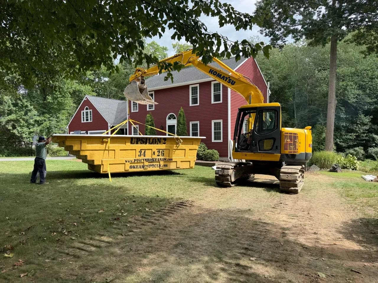 Excavator lifting an Okeanos fiberglass pool shell beside a red house.