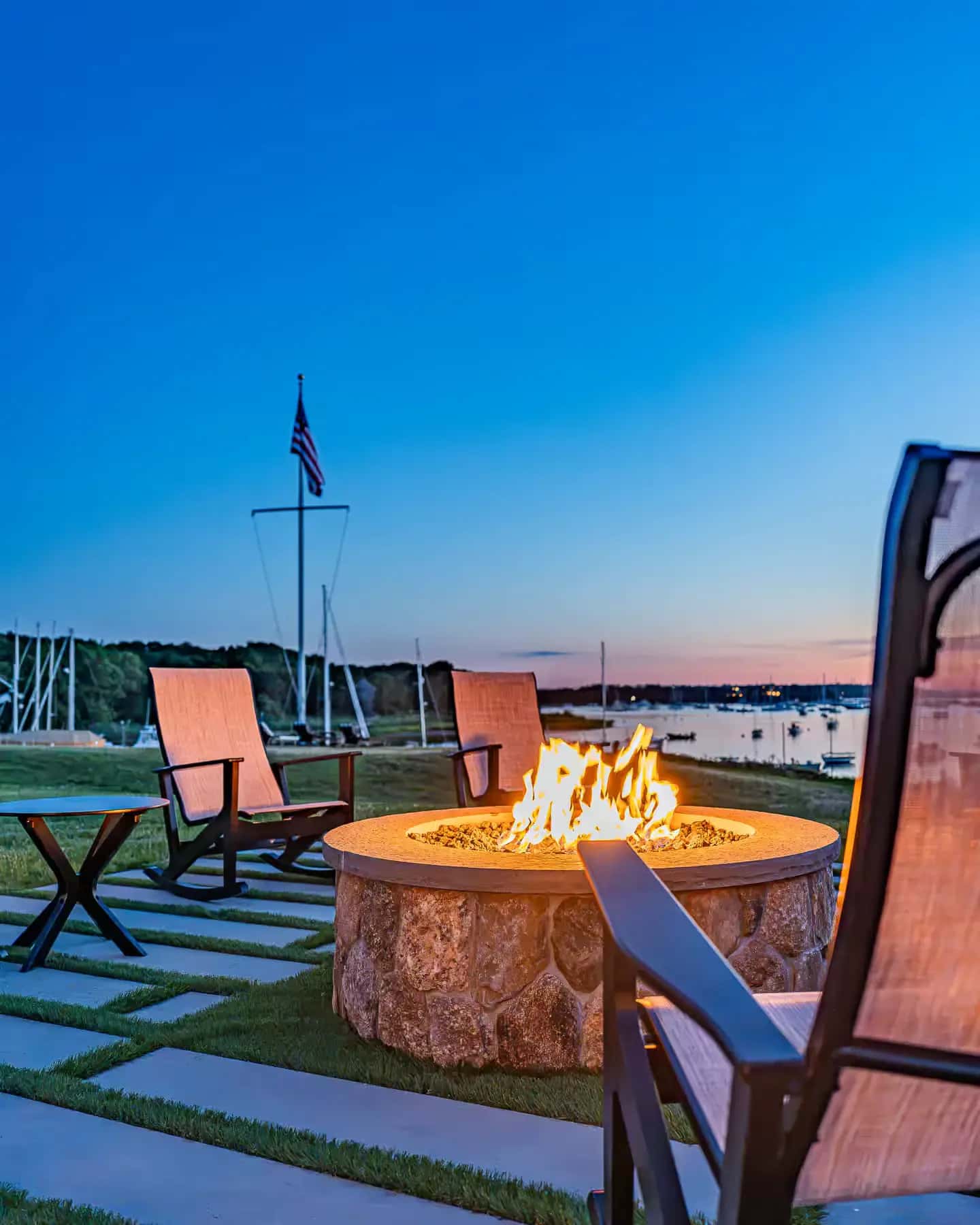 Round stone fire pit and Adirondack chairs on a grass-jointed paver terrace by the water in Buzzards Bay, MA