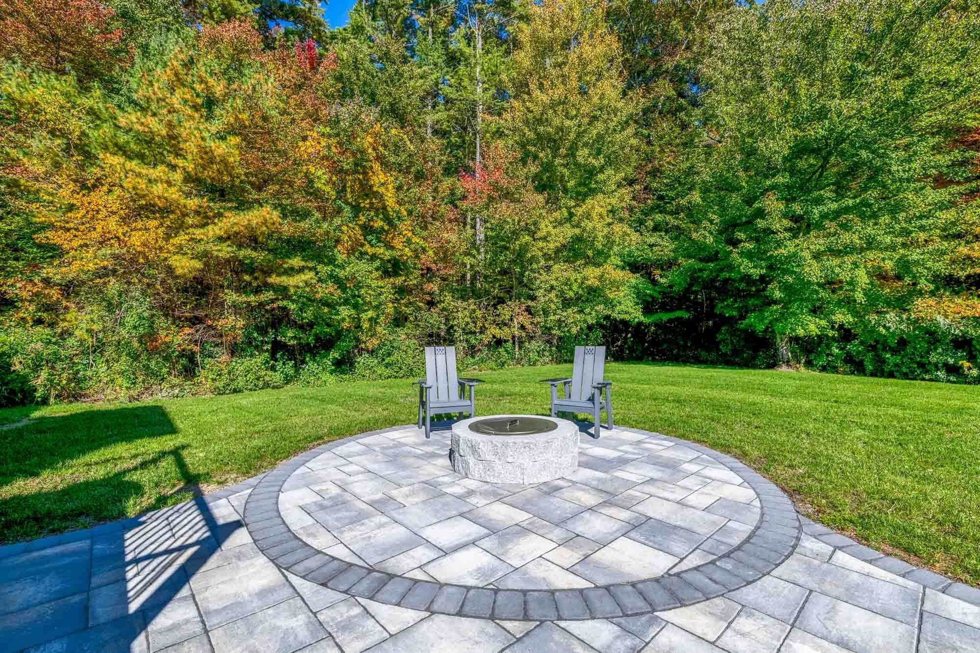 Circular paver fire-pit area with a dark border and two chairs at the lawn’s edge.
