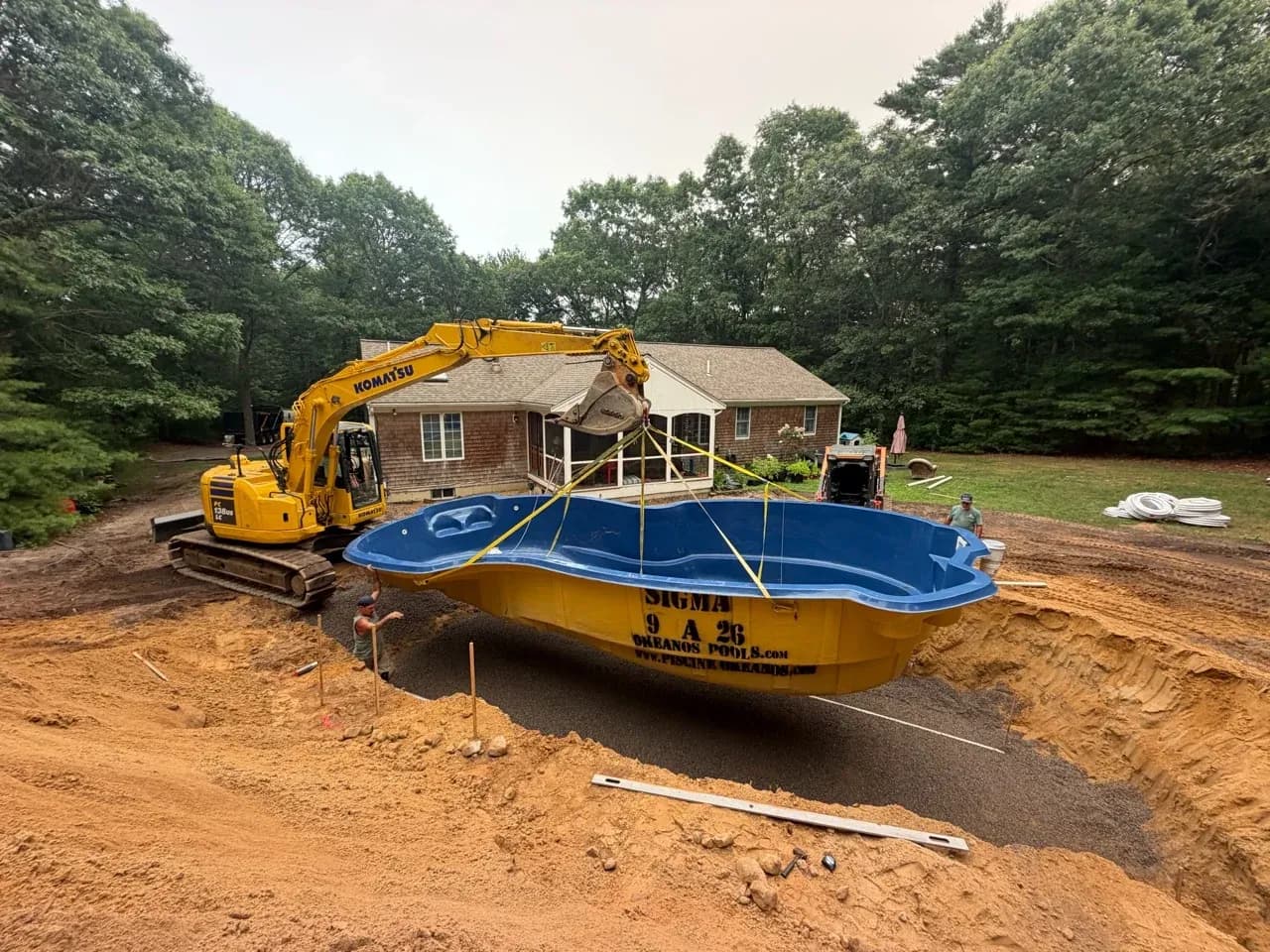 Excavator lowering a blue Okeanos fiberglass pool shell toward a prepared gravel base.