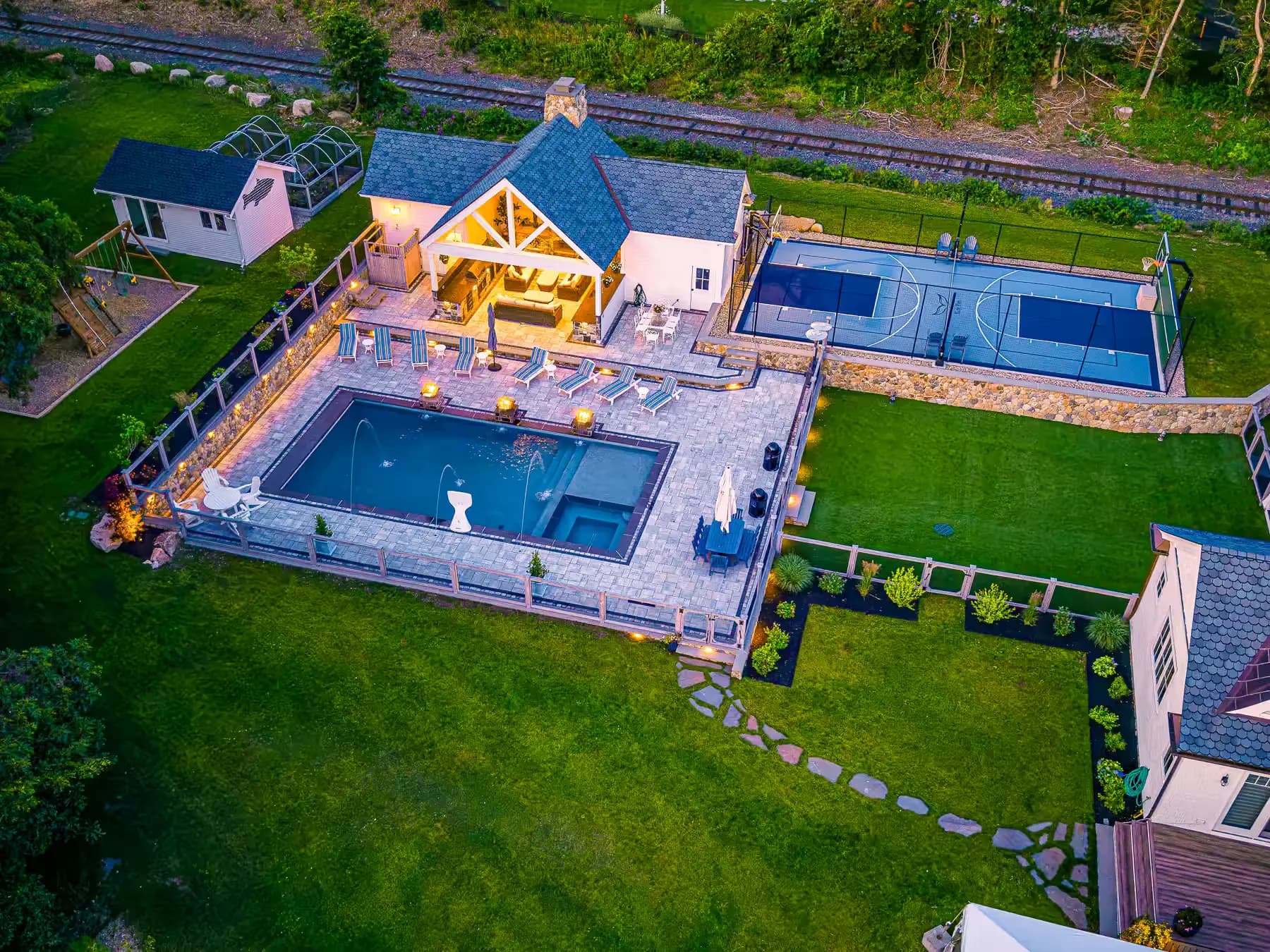 Evening aerial view of the illuminated pool, covered pavilion, sport court, and stepping-stone lawn path.