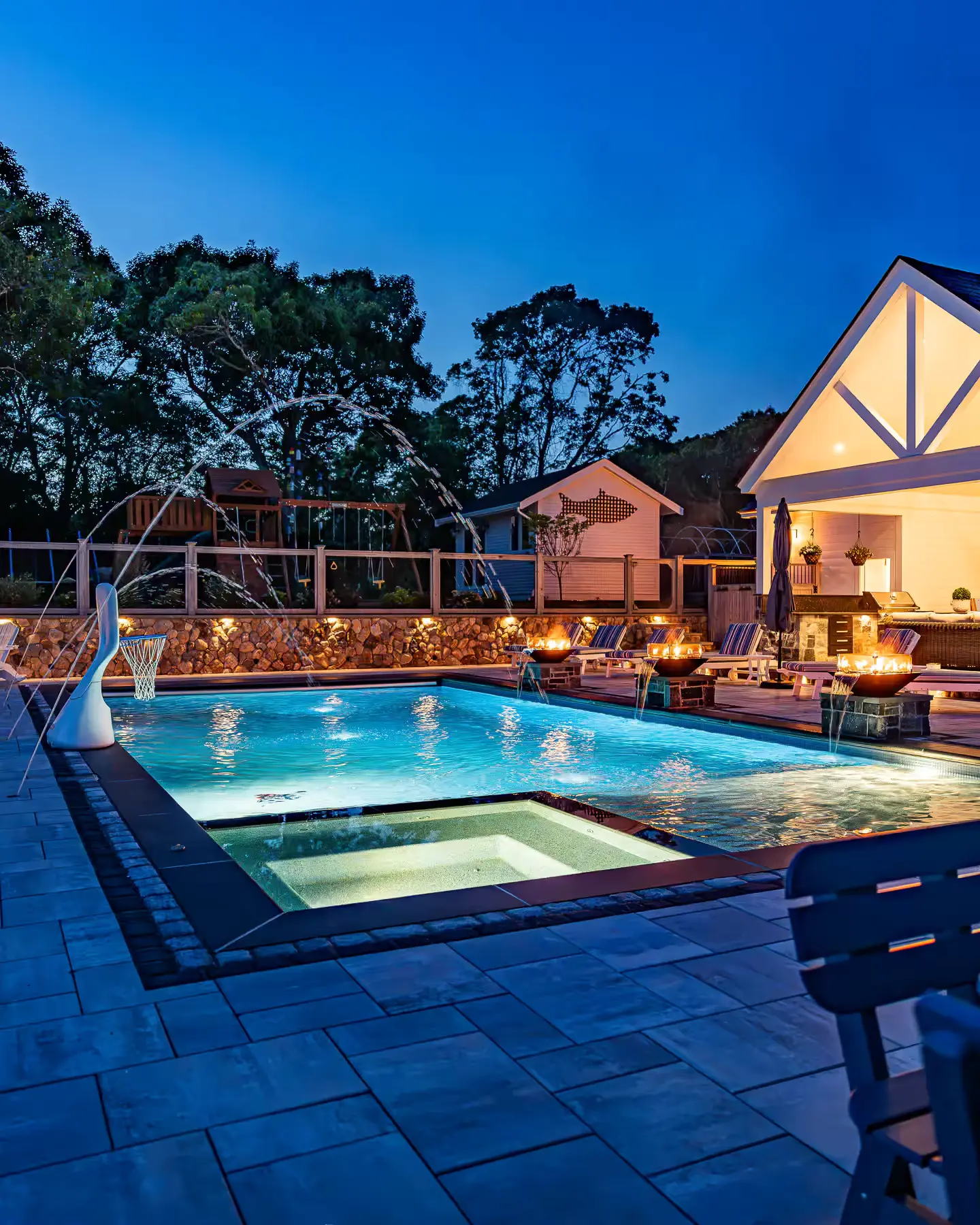 Illuminated pool and spa with fountains, fire bowls, and pavilion seating after sunset.